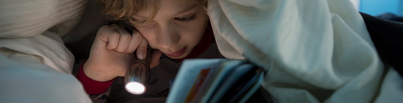 Little boy hiding under covers with flashlight reading a book