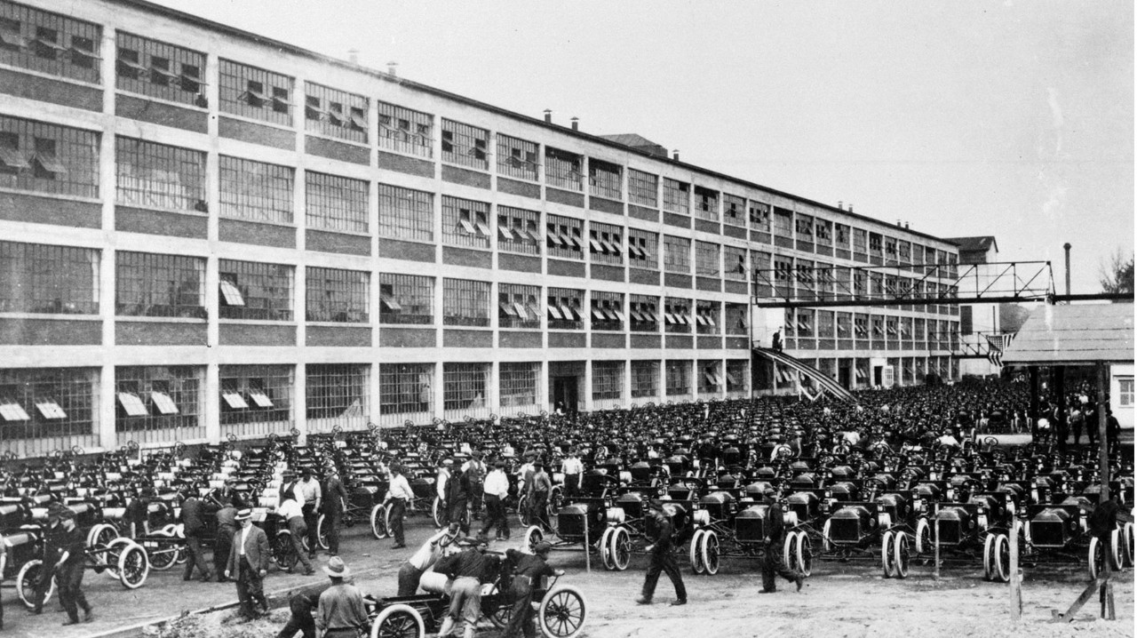 A black and white photo of hundreds of automobiles lined up in front of a Ford plant with many men walking around