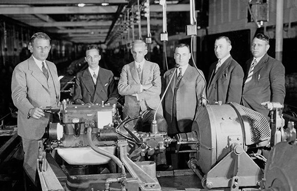 A black and white photo of Henry Ford lined up with executives inspecting a Model A engine 
