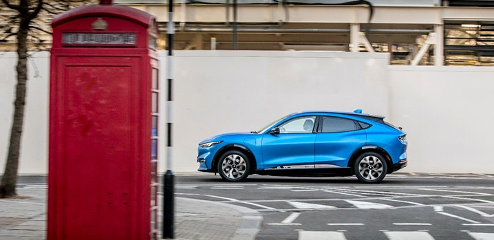 A blue Mustang MachE driving down a street near a red phone booth